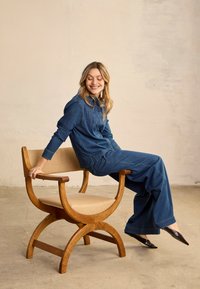 Femme en tenue en jean assise de côté sur une chaise en bois avec un rembourrage beige, souriante et regardant vers le bas, dans une pièce austère.