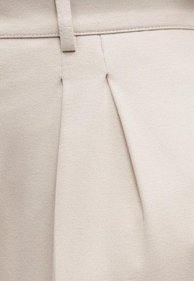 Κοντινό πλάνο μπεζ ύφασματος με πιέτα και θηλιά ζώνης, που δείχνει τις λεπτομερείς ραφές και την υφή.