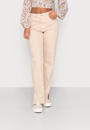 Femme portant un pantalon beige taille haute à jambes larges, un top court fleuri à manches longues et des baskets beiges à semelles blanches.