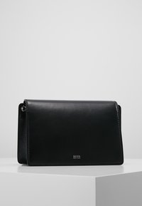 Bolso bandolera de cuero negro con cierre de solapa, con textura lisa y detalle sutil del logo en la parte inferior delantera.