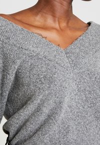 Pull en tricot gris avec un design décolleté en V dénudé, offrant une texture douce et une coupe ample, mettant en valeur le cou et les épaules.