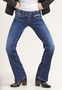 En person klädd i mörkblå utsvängda jeans, svarta klackboots, en vit skjorta och en svart läderjacka, står med benen isär och händerna på höfterna.