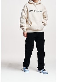 Krémová mikina s nápisem "2Y STUDIOS" černým písmem, v kombinaci s černými cargo kalhotami se zásuvnými kapsami a modro-bílými teniskami.
