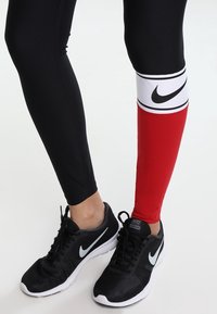 Leggings de Nike con una tela negra y una sección acentuada en rojo y blanco, combinados con zapatillas negras con un swoosh blanco.