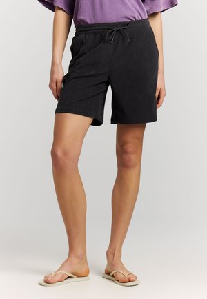 Persoon die zwarte shorts met trekkoord, paarse T-shirt en beige slippers draagt, staand tegen een effen lichte achtergrond.