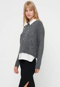 Pulover gri cu accente de perle, stratificat peste o cămașă albă cu guler. Tricotul texturat are mâneci lungi și un design scurt.