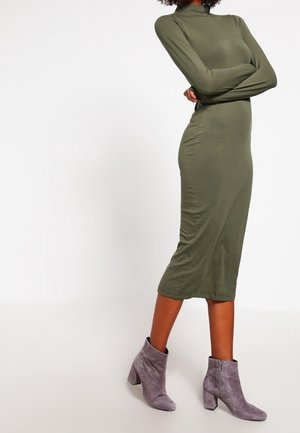 Femme portant une robe midi vert olive à manches longues et des bottines en daim grises, debout, bras croisés devant un fond blanc.
