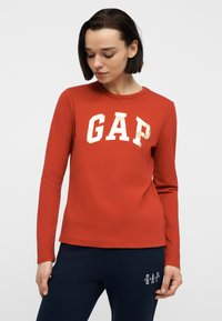 Γυναίκα που φοράει μακρυμάνικο πουκάμισο σε σκουριά χρώμα με λαμπερά γράμματα "GAP" και σκούρο μπλε παντελόνι με λευκό λογότυπο "GAP" στον μηρό.