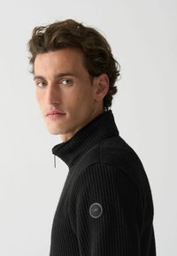 Jauns vīrietis ar cirtainiem brūniem matiem, ģērbies melnā ribotā zip-up džemperī ar Colmar logo uz pleca, skatās šķībi.