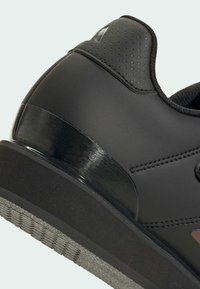 Scarpa sportiva nera con una finitura opaca liscia, tallone testurizzato e un accento lucido. Presenta una suola in gomma con un battistrada pattern.