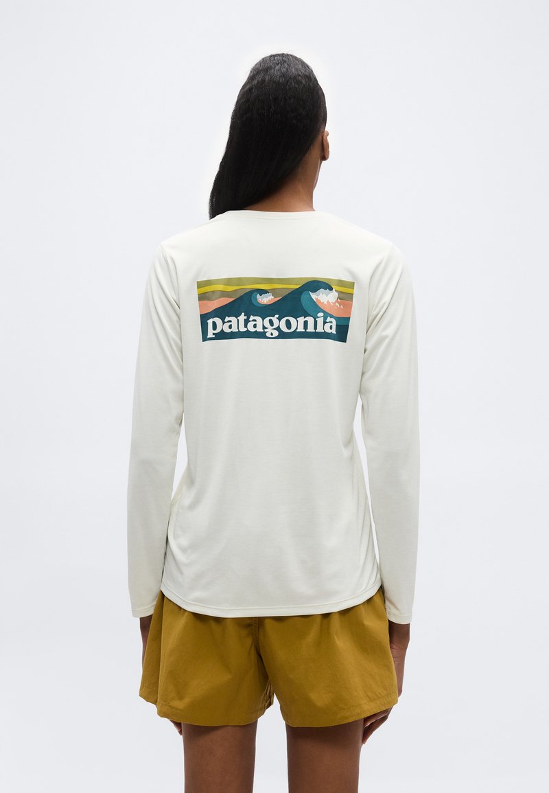 Άτομο που φοράει λευκό μακρυμάνικο πουκάμισο με το λογότυπο της Patagonia που απεικονίζει στιλιζαρισμένα ωκεάνια κύματα, συνδυασμένο με μουσταρδί κίτρινα σορτς, στέκεται μπροστά σε λευκό φόντο.