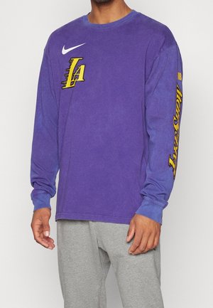 Mann trägt ein lila Langarmshirt mit gelbem Los Angeles Lakers-Logo und Nike-Swoosh, kombiniert mit hellgrauen Hosen.