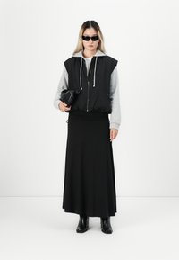 Vrouw met zwarte zonnebril, mouwloos zwart zip vest over een grijze hoodie, lange zwarte rok, zwarte laarzen, terwijl ze een zwarte clutch vasthoudt tegen een witte achtergrond.