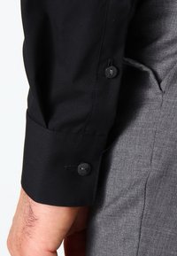 Mouw van een zwarte blouse met knoopjes die rust op de zak van grijs gestructureerde broeken op de arm van een persoon.