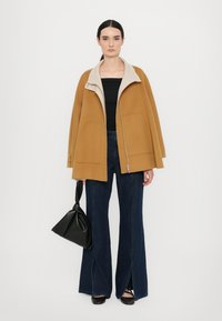 Cappotto di lana color cammello con colletto beige a contrasto, dotato di due tasche e chiusura con zip, abbinato a jeans scampanati scuri e una borsa nera.