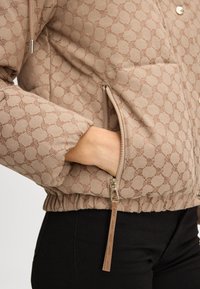 Beżowa pikowana kurtka z powtarzającym się wzorem, z zamykaną na zamek kieszenią, elastycznym brzegiem i gładką fakturą. Akcenty w kolorze srebrnym.