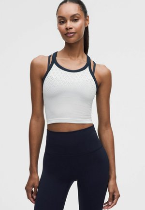 Mujer con top deportivo blanco sin mangas y corto con ribete negro y leggings negros de talle alto sobre un fondo liso.