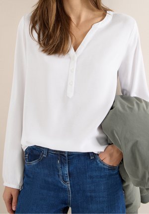 Femme portant un t-shirt henley blanc à manches longues et un jean bleu, tenant une veste grise-verte pliée, une main dans la poche.