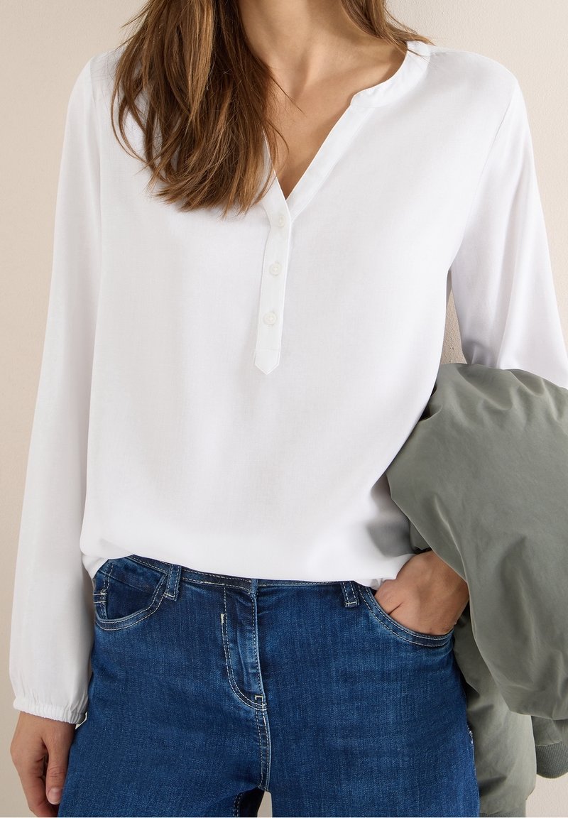 Femme portant un t-shirt henley blanc à manches longues et un jean bleu, tenant une veste grise-verte pliée, une main dans la poche.