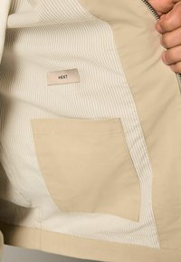 L'intérieur de la veste beige présente un tissu lisse avec un motif rayé, une petite poche cousue et une étiquette indiquant "NEXT."