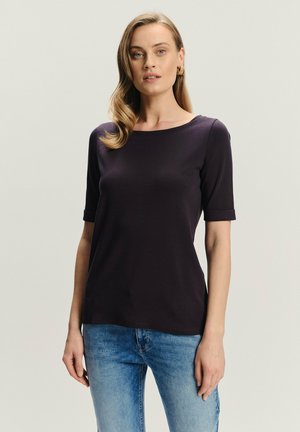 Kobieta z falującymi blond włosami, nosząca złote kolczyki w kształcie obręczy, ciemnofioletową bluzkę z krótkim rękawem i jasnoniebieskie dżinsy, stojąca na jednolitym tle.