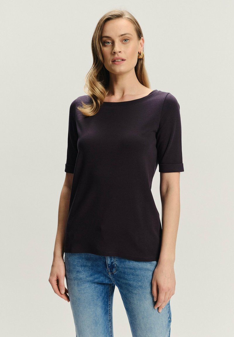 Kobieta z falującymi blond włosami, nosząca złote kolczyki w kształcie obręczy, ciemnofioletową bluzkę z krótkim rękawem i jasnoniebieskie dżinsy, stojąca na jednolitym tle.