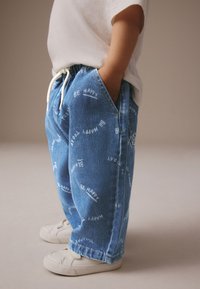 Niebieskie spodnie z denimu z białymi napisami "BE HAPPY" i "HAPPY TO" oraz elastycznym pasem, zestawione z beżowymi sneakersami.