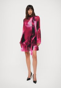 Mulher vestindo um vestido floral rosa e preto, de manga longa e comprimento até os joelhos, com um laço no pescoço, combinado com sapatos de salto alto pretos, em pé sobre fundo branco.