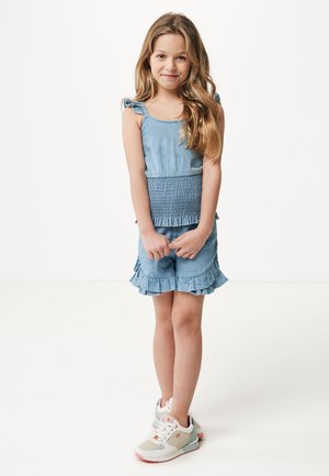 Vestido de mezclilla azul claro con cuerpo fruncido, dobladillo con volantes y tirantes. Combinado con zapatillas blancas y en tonos pastel.