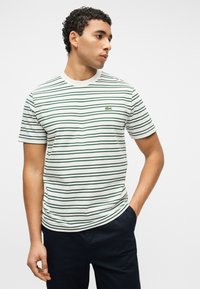 Άνδρας με κοντά σγουρά μαλλιά που φοράει λευκό και πράσινο ριγέ κοντομάνικο πουκάμισο και σκούρο παντελόνι, στέκεται με το ένα χέρι στην τσέπη.