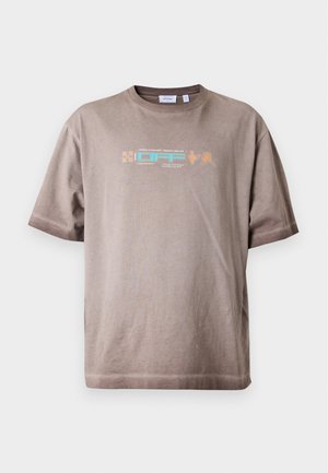 Jeune homme debout portant un t-shirt graphique marron clair, un pantalon ample foncé, des baskets blanches, une main dans sa poche, devant un fond uni.