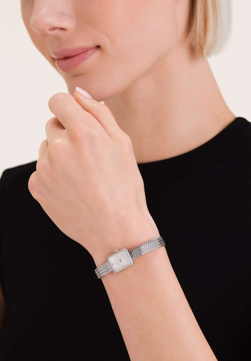 Montre bracelet en maille argentée avec un cadran carré blanc, affichant un design minimaliste et des accents subtils. Portée sur un teint clair.