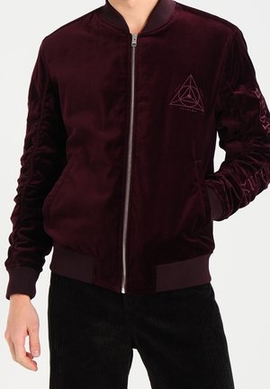 Burgunderfarbener Samt-Bomberjacke mit gerippten Bündchen, Reißverschluss und einem geometrischen Logo auf der linken Brust. Eine Cordhose vervollständigt den Look.