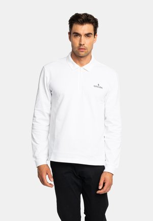 Mann trägt ein weißes, langärmeliges Poloshirt mit "Gisada"-Logo auf der linken Brust und schwarze Hose, steht vor einfarbigem Hintergrund.