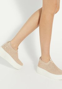 Beige knitted slip-on sneakers with a thick white sole, featuring a ribbed collar and a textured upper with subtle sparkle details.