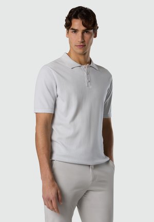 Giovane uomo in piedi con le mani in tasca, indossa una polo bianca a maniche corte e pantaloni beige chiaro su uno sfondo neutro.