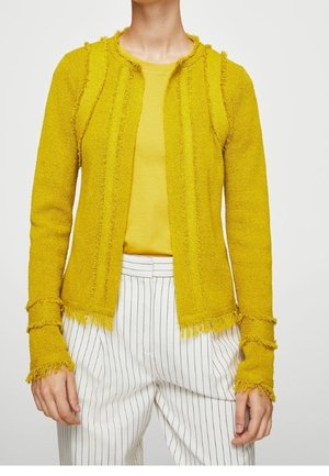 Kobieta w musztardowożółtej, teksturowanej kurtce z frędzlami, w pasującej żółtej koszuli i białych spodniach w cienkie paski.