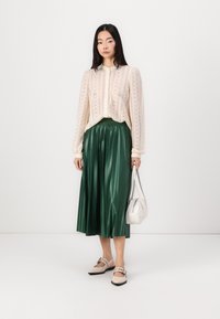 Top de encaje crema con mangas largas y un escote alto, combinado con una falda de satén verde plisada y zapatos beige, sosteniendo un pequeño bolso blanco.