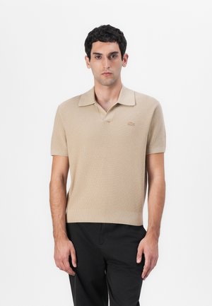 Polo beige à manches courtes avec une texture côtelée, un col plat et un logo brodé subtil sur la poitrine. Associé à un pantalon sombre.