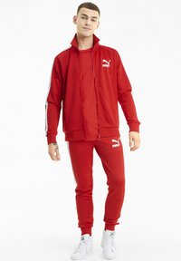 Ensemble de sport rouge en tissu lisse, comprenant une veste zippée avec des accents blancs et un pantalon assorti, portant tous deux le logo Puma.