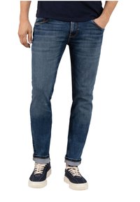 Blaue Denim-Jeans im Slim Fit, mit leichten Used-Effekten und umgeschlagenen Säumen, kombiniert mit dunklen Sneakers mit weißen Sohlen und Schnürsenkeln.
