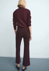 Persoană cu păr blond scurt, purtând un pulover burgund închis și pantaloni evazați asortați, stând pe covor gri.