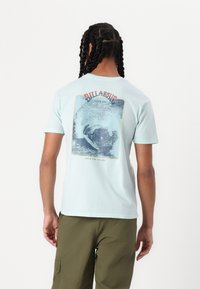 Lyseblå bomull t-skjorte med korte ermer, med et grafisk motiv av en bølge på ryggen og teksten "Billabong Siden 1973" over.