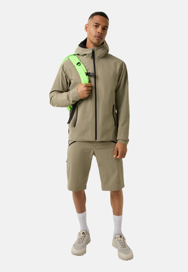 Beige waterproof jacket with black zippers, paired with matching shorts. Features a bright green backpack and gray athletic shoes with white accents.
