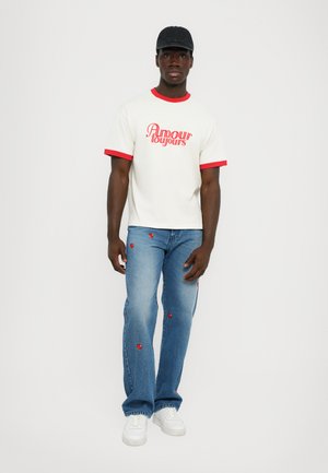 White t-shirt with red trim and text, paired with blue jeans featuring red heart embroidery, and white sneakers. Man stands upright.