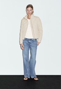 Beige gewatteerde jas met een ritsluiting, gestructureerd oppervlak en hoge kraag, gecombineerd met lichtblauwe wijde Jeans en zwarte sandalen.