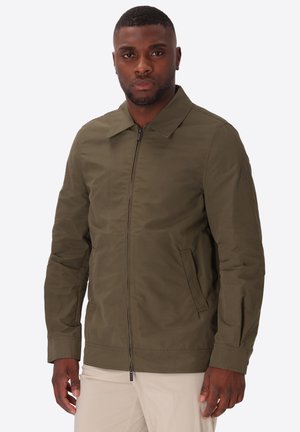 Uomo che indossa una giacca verde oliva con zip, colletto e tasche laterali, in piedi davanti a uno sfondo chiaro e semplice.