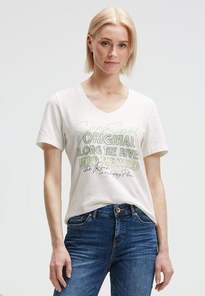 Young woman with short blonde hair wearing a white V-neck T-shirt with multicolored text and blue jeans, standing against a plain background.