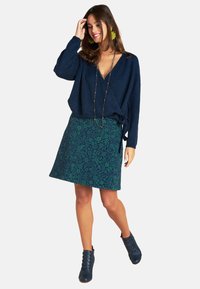 Dunkelblaue, gestrickte Wickeljacke mit langen Ärmeln, kombiniert mit einem grünen, floralen A-linien Rock und blauen Stiefeletten.
