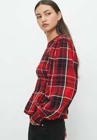 Chemise à carreaux rouge et noir avec des manches bouffantes, un corsage smocké et un col rond. Présente un tissu texturé et une coupe ajustée à la taille.